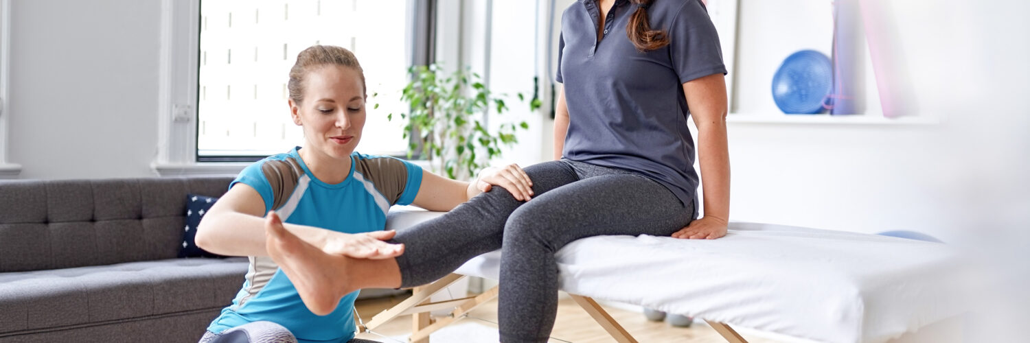Naisfysioterapeutti on lattialla kyykyssä ja naisasiakas ojentaa jalkaansa suoraksi eteen.
