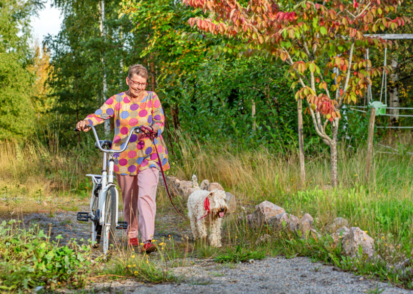 Iäkäs nainen taluttaa pyörää koiran kanssa ruska-aikaan.