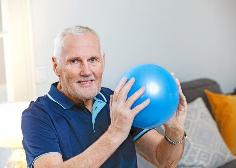 Seniorimies pitää sinistä jumppapalloa käsissä ja katsoo kohti.