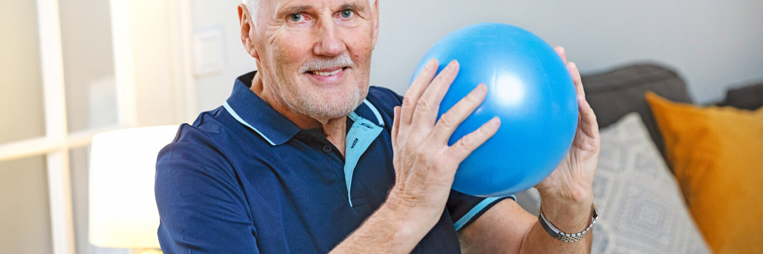 Seniorimies pitää sinistä jumppapalloa käsissä ja katsoo kohti.
